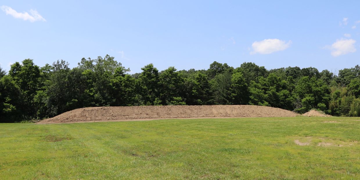Large mound of dirt in a green field, with trees and blue sky in the background.