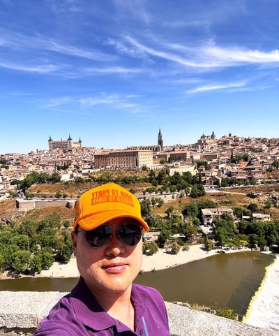 Man wearing orange cap and sunglasses taking a selfie with a historic cityscape and river in the background.