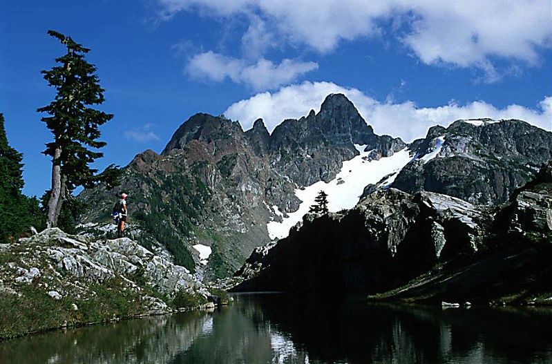 A lone hiker stands near a mountain lake beneath rugged snowy peaks.