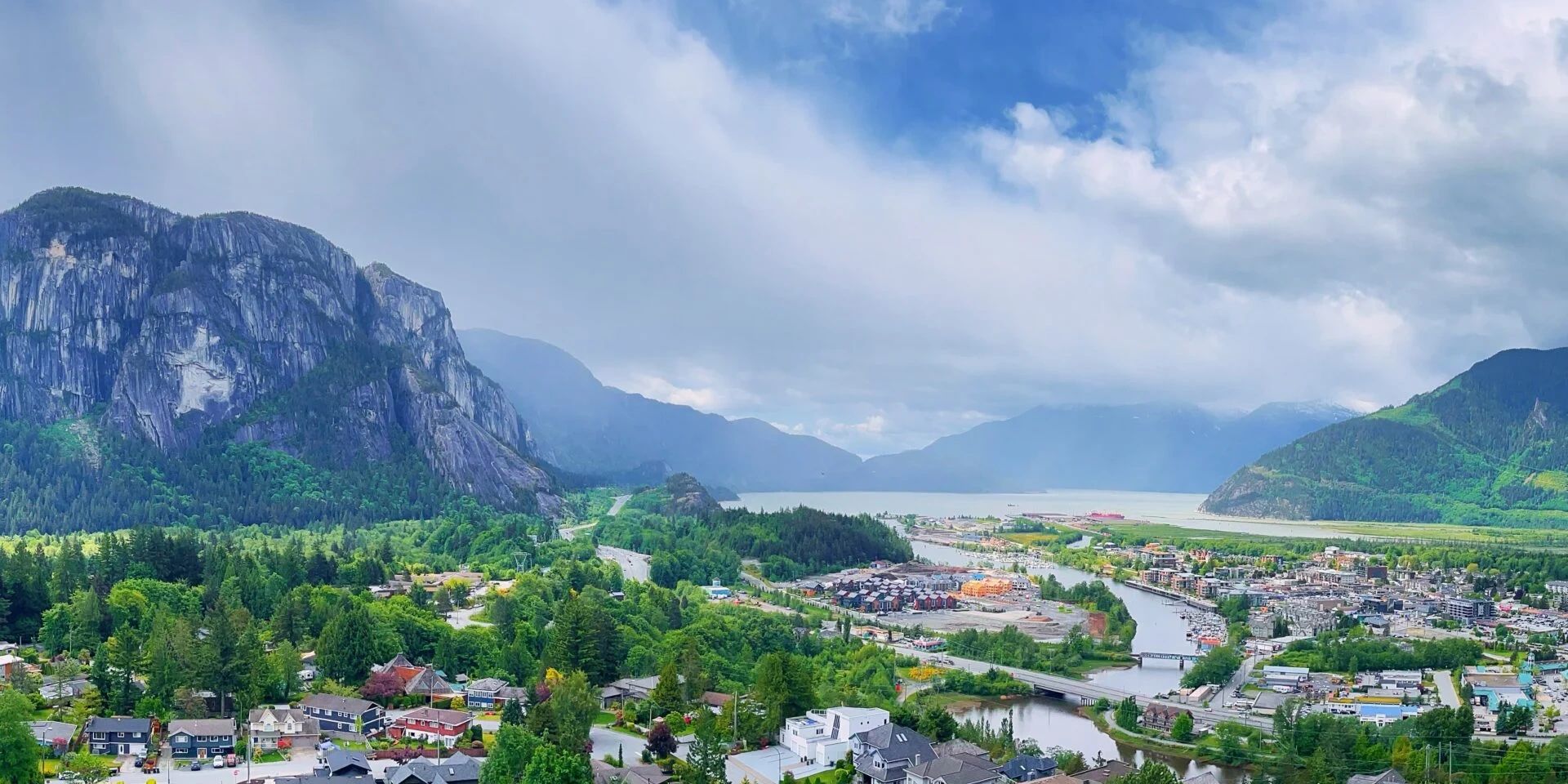 A scenic town nestled between mountains and a river under a partly cloudy sky.