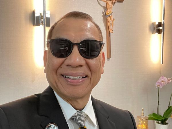 A man in a suit wearing sunglasses smiles indoors with a crucifix and flowers behind him.