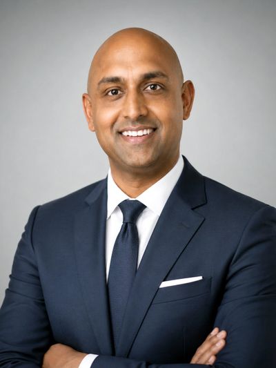 Confident man in a dark suit smiling with arms crossed against a gray background.