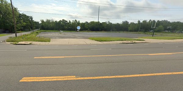 Empty parking lot beside a road with trees in the background.