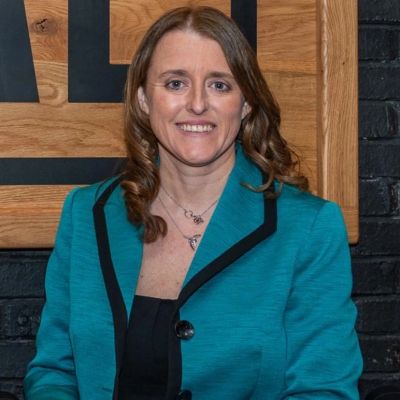 Woman in teal blazer smiling against wooden and black background.