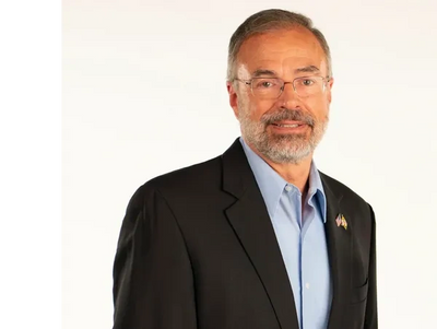 Man wearing glasses and a dark suit with a light blue shirt, smiling.