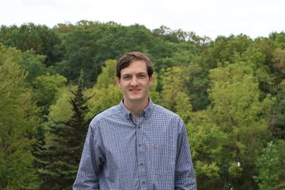 A man in a checkered shirt stands outdoors with green trees in the background.