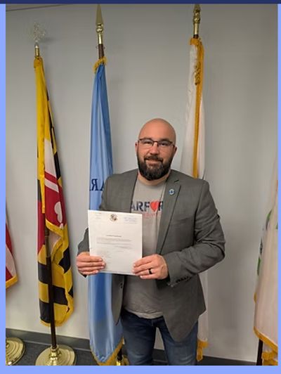 Man in blazer holding a document, standing between flags indoors.