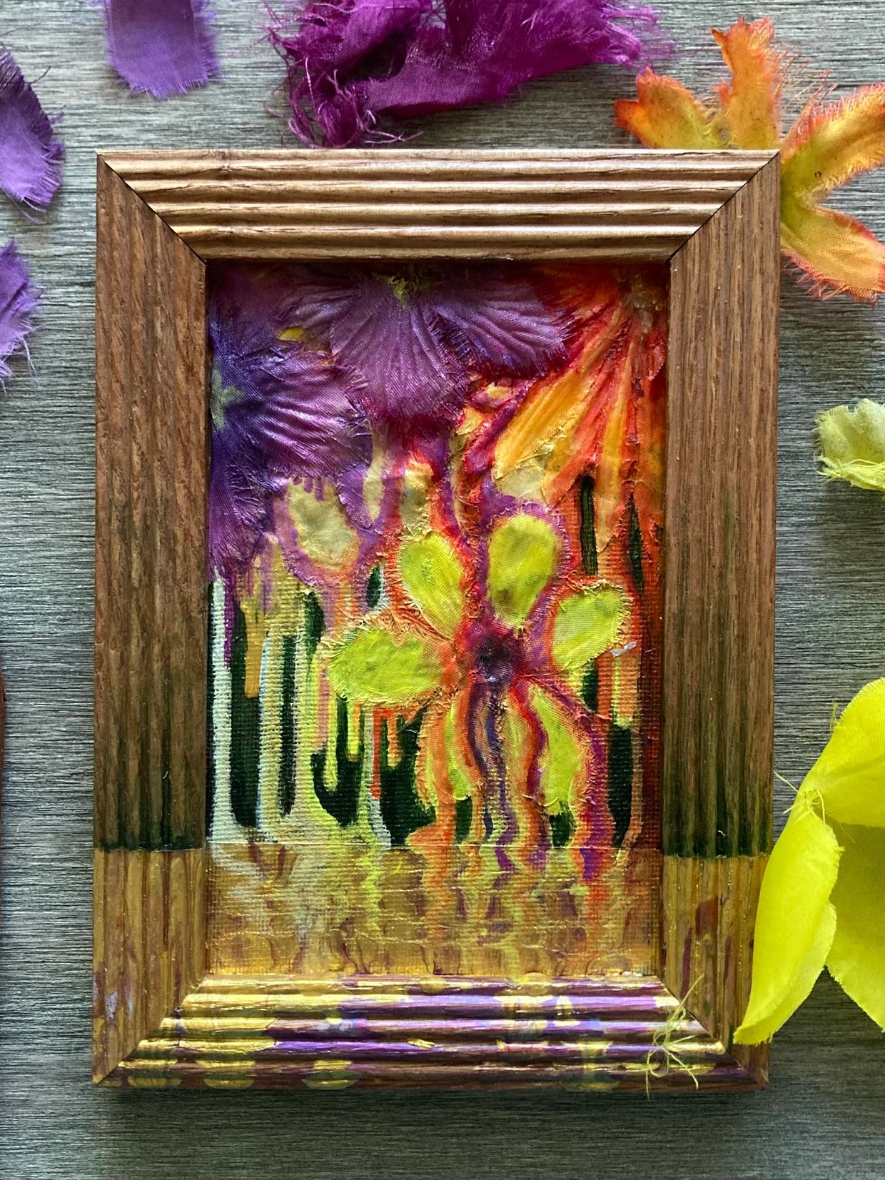 Framed fabric art with vibrant purple, orange, and yellow flowers on a wooden surface.