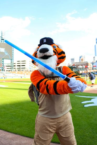 Detroit Tigers mascot holding a blue lightsaber on a baseball field.