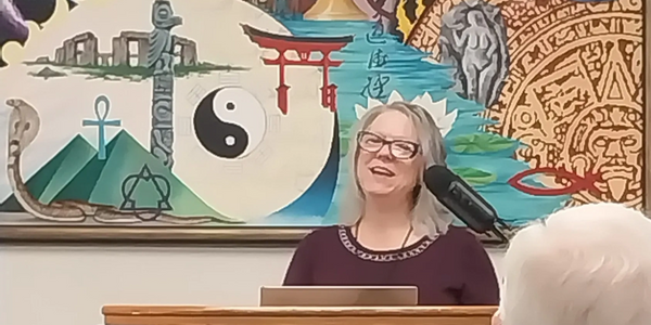 White, woman, w/brown hair & glasses preaching in front of mural depicting many different religions