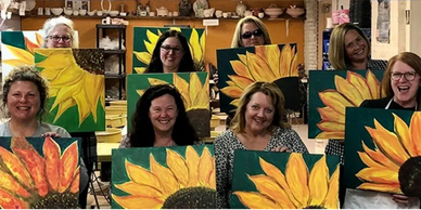 Group of women proudly displaying their sunflower paintings at a painting class.
