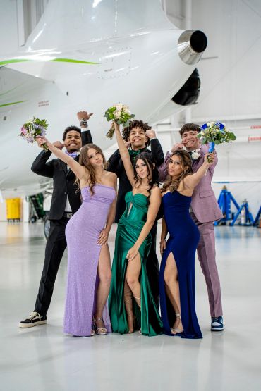 Group of young people in formal attire holding bouquets, posing cheerfully in front of a plane.