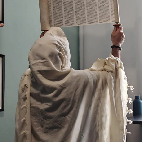 A person wrapped in a tallit holding up a Torah scroll indoors.