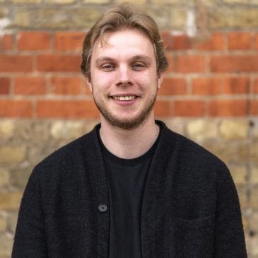 Smiling man in a black cardigan stands against a brick wall backdrop.