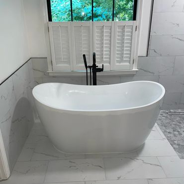 Modern white freestanding bathtub in a bright bathroom with window shutters.