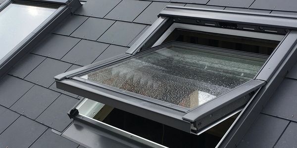 Open skylight window on a dark tiled roof with raindrops.