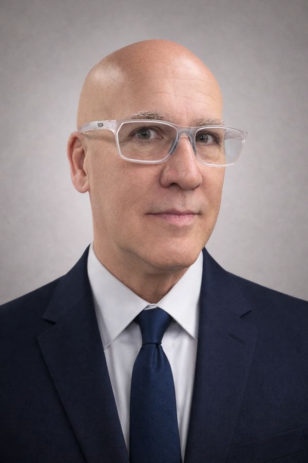 Professional bald man wearing clear glasses and a navy suit.