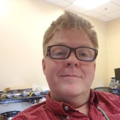 Smiling man with glasses in a red shirt at an office desk.
