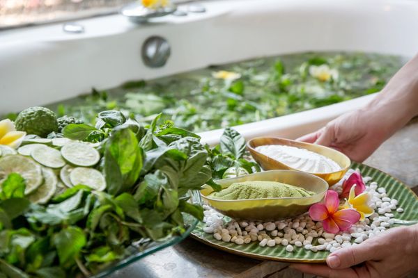 Hands hold a tray with bowls of green powder and white granules next to a herbal bath