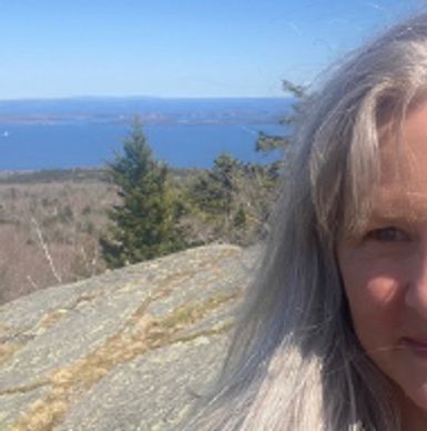 Woman with long gray hair outdoors near rocks and trees with ocean in the background.