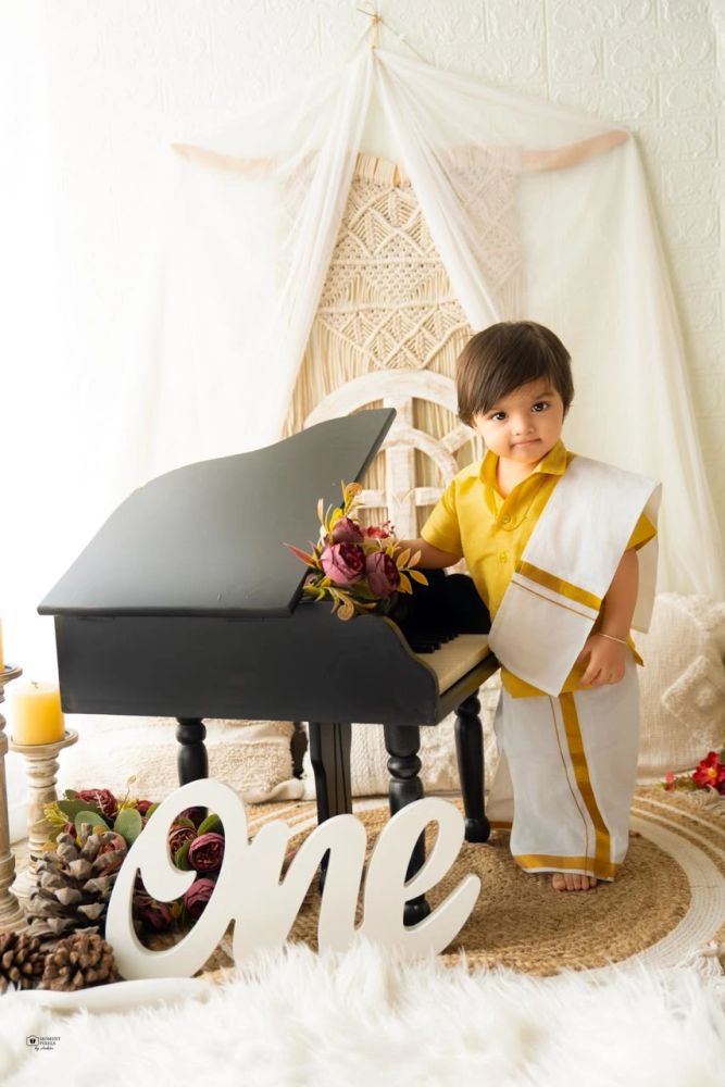 Toddler boy in traditional attire by a small piano with 'One' sign.
