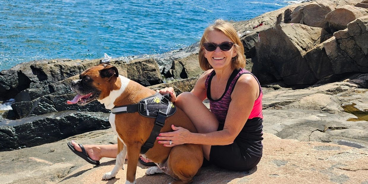 Woman in sunglasses sitting on rocks by the sea with her dog.