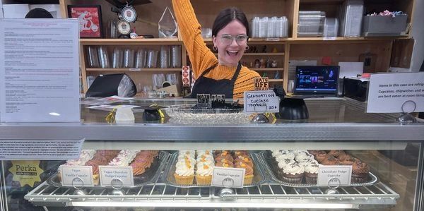 One of our employees posing behind the counter with a variety of cupcakes in the case