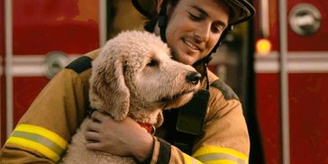 Firefighter warmly hugging a fluffy dog in front of a fire truck.