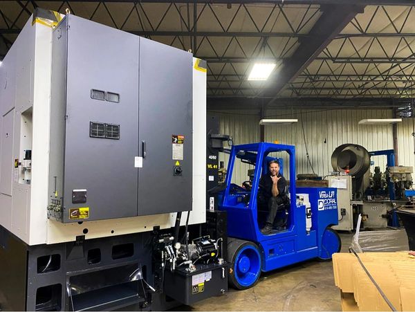 Man sitting on a blue forklift inside an industrial workshop.