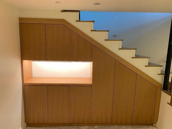 Wooden storage cabinets built under a staircase with a lit open shelf.