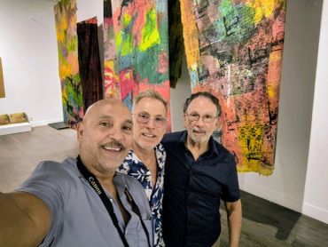 Three men posing in an art gallery with colorful abstract paintings.