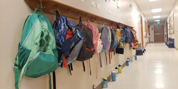 Colorful backpacks hanging on hooks in a school hallway with water bottles lined up below.