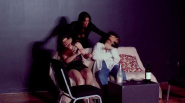 Three people interacting in a dark room with chairs and a table.