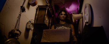 Woman sitting in a dim, cluttered room lit by purple light.
