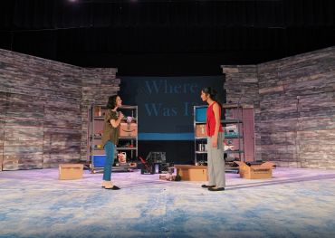 Two actors perform on a rustic stage set with boxes and shelves.