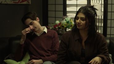 Two people sit on a couch looking thoughtful and tired.