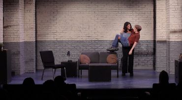 Two women acting in a theater scene with minimalist set.