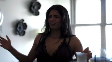 Woman in dark outfit gesturing and holding a cup indoors.