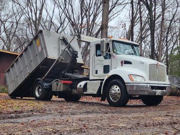 White dump truck unloading a large container in a wooded area.