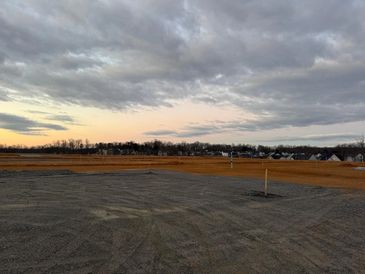 A cloudy sky over an empty gravel lot with distant houses and fields.