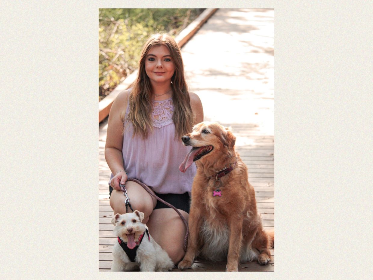 Young woman kneels with two happy dogs on a sunny wooden path.