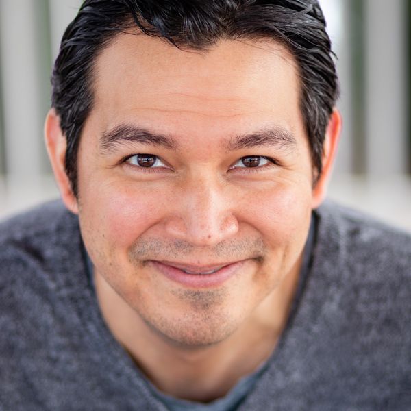 Shayne Steliga smiles at the camera in front of a white and gray striped background.