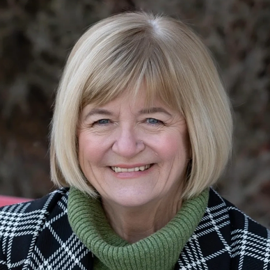 Smiling elderly woman with blonde hair wearing a green sweater and checkered coat.