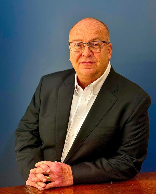 A mature man in a black suit with glasses poses confidently.