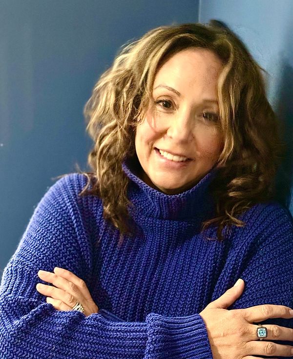 Woman smiling warmly in a cozy blue sweater with arms crossed.