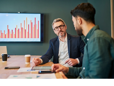 Two men having a business discussion with a bar chart displayed on a screen.