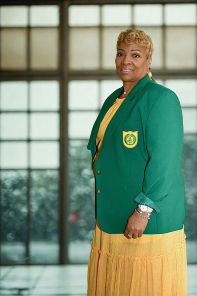 Woman in green blazer and yellow dress smiling indoors.