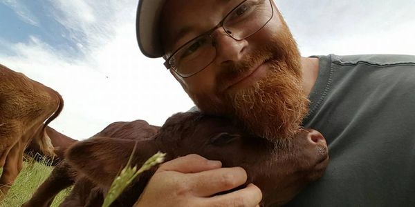 Man with glasses and cap hugging a brown calf outdoors.
