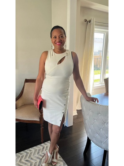 Woman in a stylish white dress holding a pink clutch, smiling indoors.