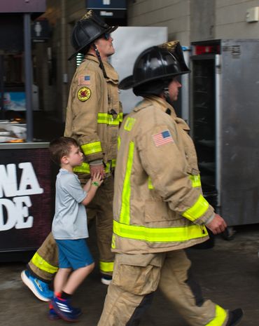 Two firefighters walk with a young boy holding one firefighter's hand indoors.